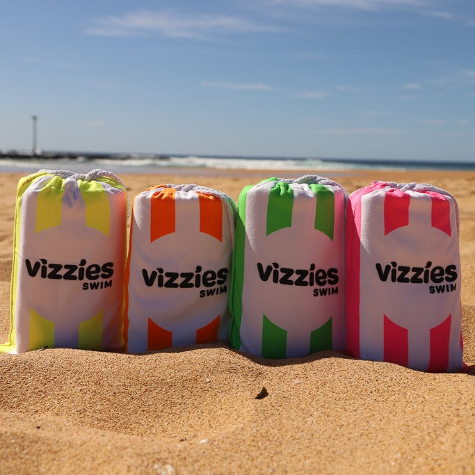 Four drawstring beach towels in white with colorful vertical stripes (yellow, orange, green, and pink) standing on a sandy beach with the ocean and blue sky in the background. The towels are branded with 'vizzies SWIM' in black text.