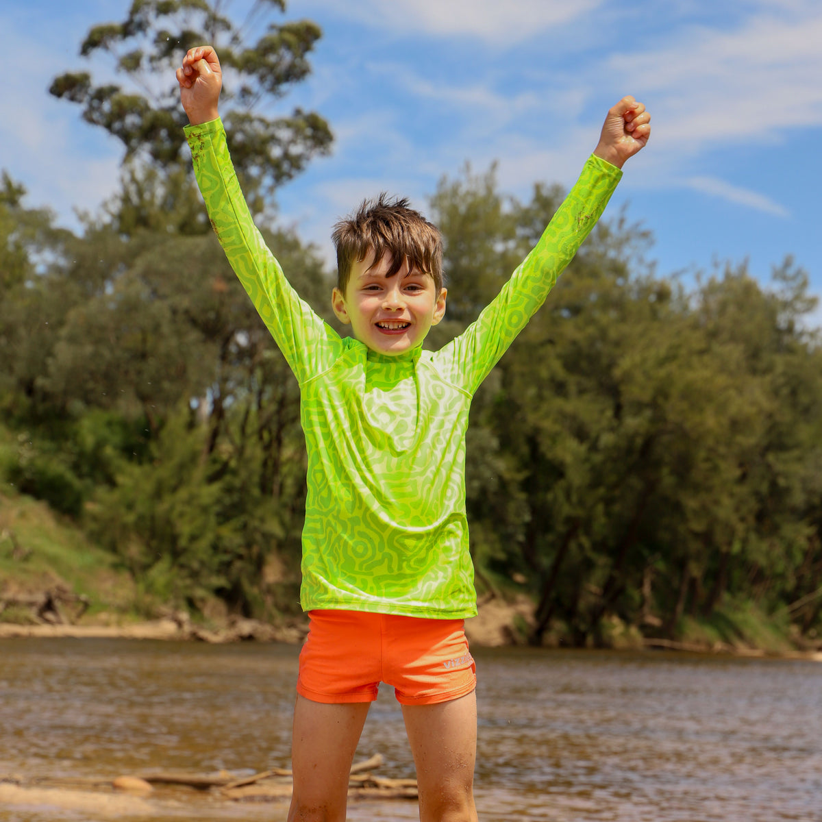 A young boy stands in front of a river with trees in the background, wearing a neon  Lime Light Print long-sleeve swim rashie and bright orange zap swimshorts from Vizzies Swim, with his arms raised in excitement.