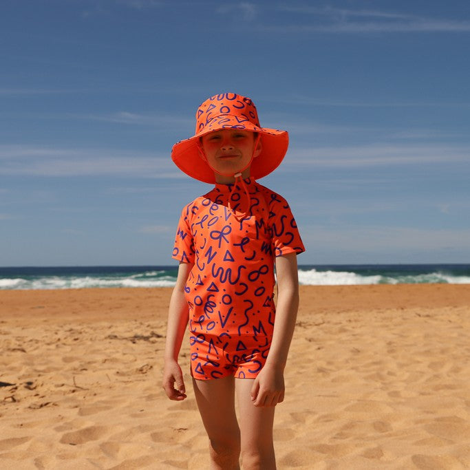 A child standing on a sandy beach wearing a bright orange rashie and swim shorts from Vizzies Swim with a navy abstract pattern and a matching wide-brim sun hat, with the ocean and blue sky in the background.