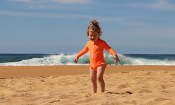 A young child wearing a bright orange long-sleeve swimsuit or rash guard stands on a sandy beach with ocean waves and a blue sky in the background.