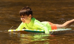A young boy wearing a neon yellow-green rash guard swims and lies on a blue bodyboard on a river or lake.