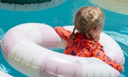 A young child wearing an orange patterned swim shirt floats on a pink and white striped inflatable pool ring in a swimming pool.