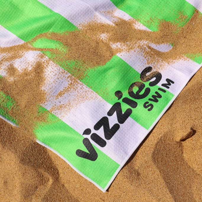 A close-up of a beach towel with green and white stripes, printed with the brand name 'Vizzies Swim' in black, lying on sand with sand scattered across it.