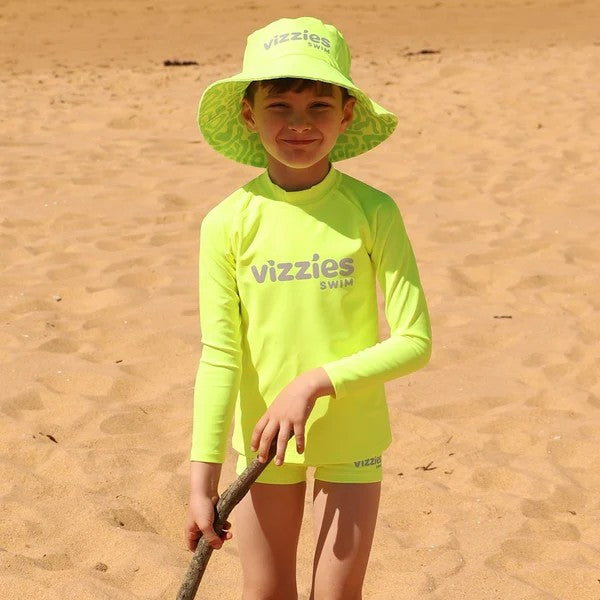 A young boy standing on a sandy beach wearing a bright neon yellow Vizzies Swim long-sleeve rashie matching swim trunks, a green patterned sun hat, and holding a stick.