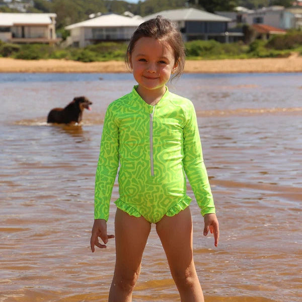 A young girl stands in shallow water at the beach wearing a bright neon green long-sleeve swim shirt with a white abstract pattern and a white front zipper from Vizzies Swim. A dog is visible in the water behind her, and houses line the distant shoreline.