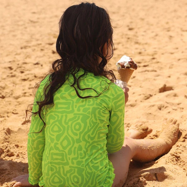 A child sitting on a sandy beach, viewed from behind, wearing a Vizzies Swim Lime Light Print long-sleeve swimsuit. The child is holding an ice cream cone.