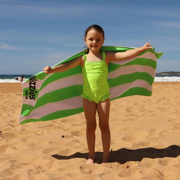 A young girl stands on a sandy beach wearing a bright neon green Vizzies swimsuit and holding a large green and white striped beach towel spread behind her like a cape. The ocean and blue sky are visible in the background.