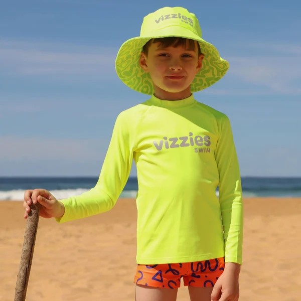 A child wearing a bright neon yellow long-sleeve rashie with 'vizzies swim' printed on the chest, a matching wide-brim sun hat, and patterned swim trunks, standing on a sandy beach with the ocean in the background.