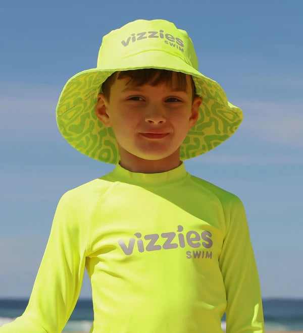 A young boy wearing a bright neon yellow-green Vizzies Swim long-sleeve rash guard and matching sun hat with a patterned brim, standing on a beach with the ocean and blue sky in the background.