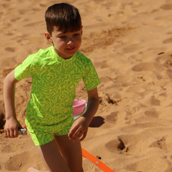 A young boy wearing a bright neon green and yellow patterned swim shirt and matching swim shorts from Vizzies Swim, standing on a sandy beach.
