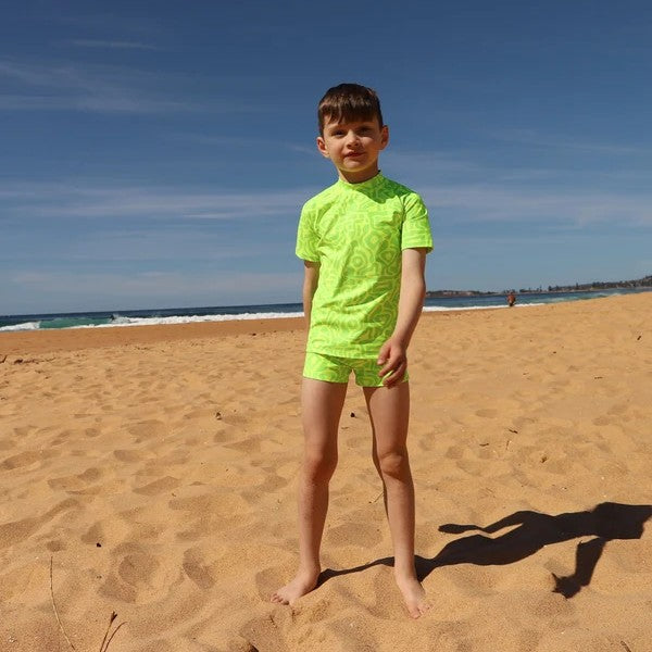 A young boy stands on a sandy beach wearing a bright neon green short-sleeve swim shirt and matching swim trunks from Vizzies Swim under a clear blue sky.