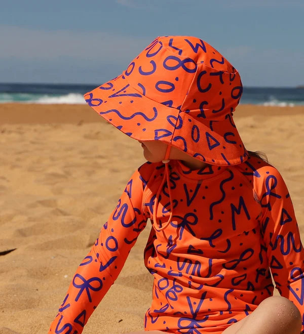 A child wearing a bright orange long-sleeve sun protection outfit with a matching sun hat from Vizzies Swim, both covered in a navy blue abstract pattern, sitting on a sandy beach with the ocean in the background.