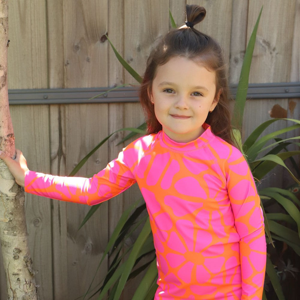 A young girl with her hair in a high ponytail smiles while wearing a long-sleeve rashie with a vibrant pink and orange abstract pattern from Vizzies Swim, standing outdoors against a wooden fence and green plants.
