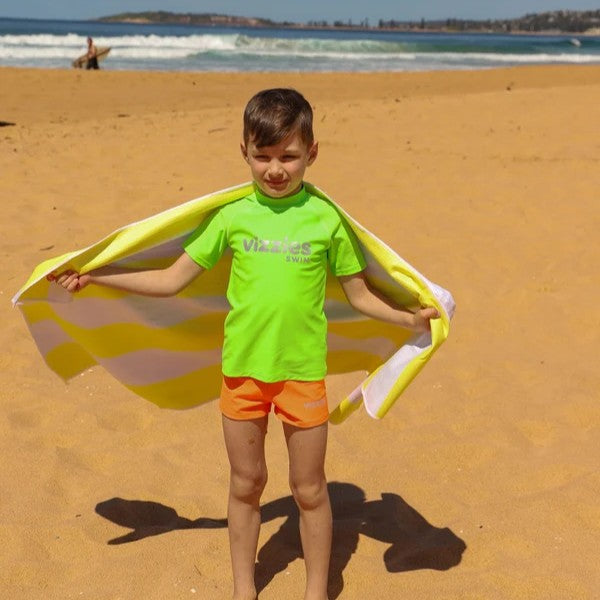 A young boy stands on a sandy beach wearing a bright neon green rash guard with 'Vizzies' printed on the chest and orange swim trunks, holding a yellow and white striped beach towel behind his back like a cape. The ocean waves and distant shoreline are visible in the background.