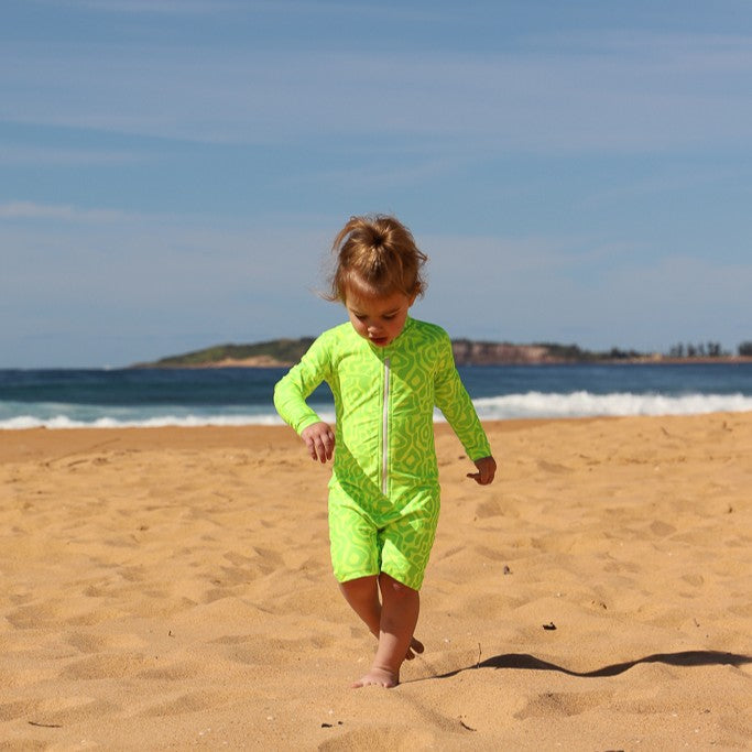 A toddler walking on a sandy beach wearing a bright neon green one-piece swimsuit from Vizzies Swim with a lighter green abstract pattern.