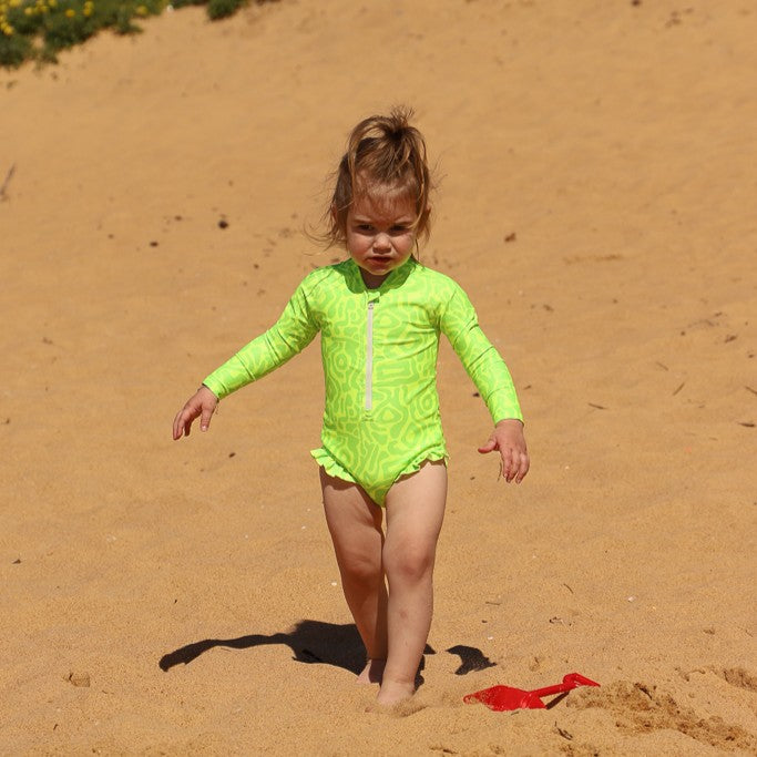 A baby walking barefoot on sandy ground wearing a bright lime green long-sleeve swimsuit with an abstract pattern on it from Vizzies Swim with  a white front zipper and frill details on the legs.