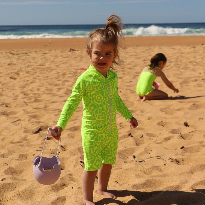 A baby wearing a bright neon green long-sleeve swimsuit with a darker green abstract pattern from Vizzies Swim stands on a sandy beach holding a small lavender bucket. Another child in a matching green swimsuit is visible in the background near the ocean.