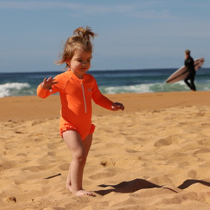 A toddler girl wearing a bright orange long-sleeve swimsuit with ruffled leg details from Vizzies Swim stands on a sandy beach with the ocean and sky in the background. A surfer is visible in the distant background.