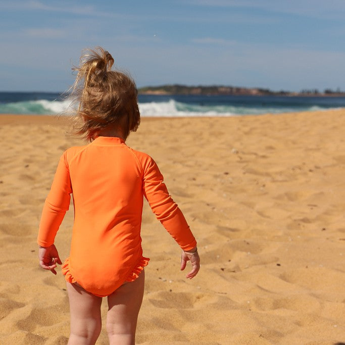 A toddler girl seen from behind walking on a sandy beach wearing a bright orange long-sleeve swim bodysuit from Vizzies Swim.