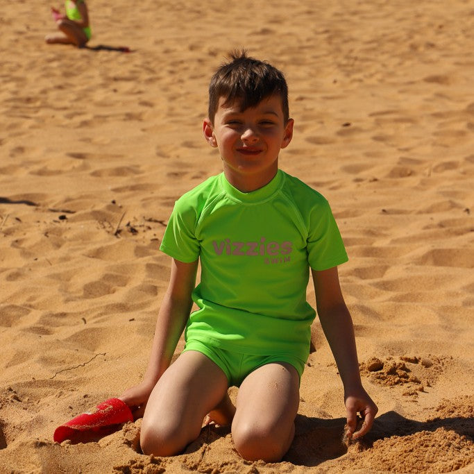 A young boy kneeling on a sandy beach wearing a bright lime green rashie and matching swim trunks, with a red toy in his hand. The brand name 'Vizzies Swim' is printed across the chest.