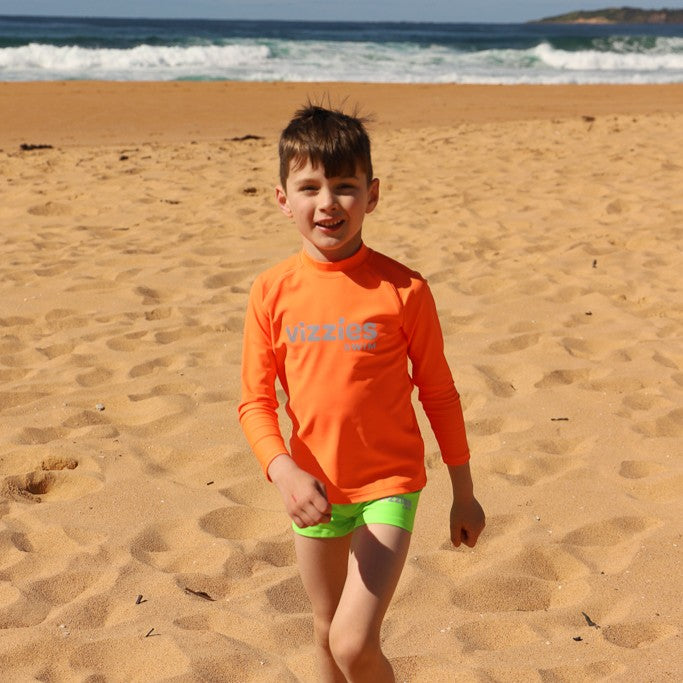 A young boy standing on a sandy beach wearing a bright orange long-sleeve swim rashie with 'Vizzies Swim' printed on the chest and neon green swim trunks, with the ocean waves in the background.