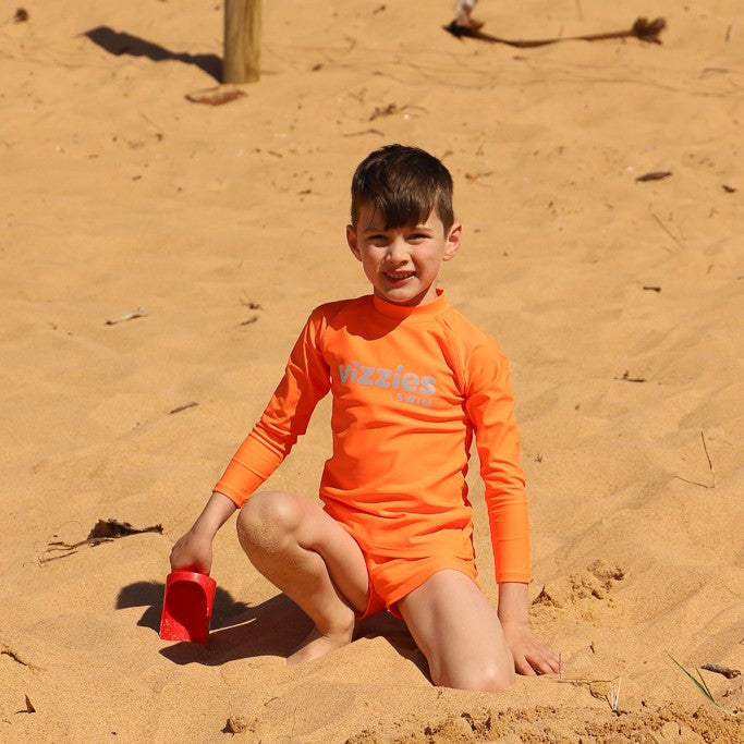 A young boy sitting on a sandy beach wearing a bright orange long-sleeve rashie and matching swim trunks from Vizzies Swim, holding a small red shovel. 