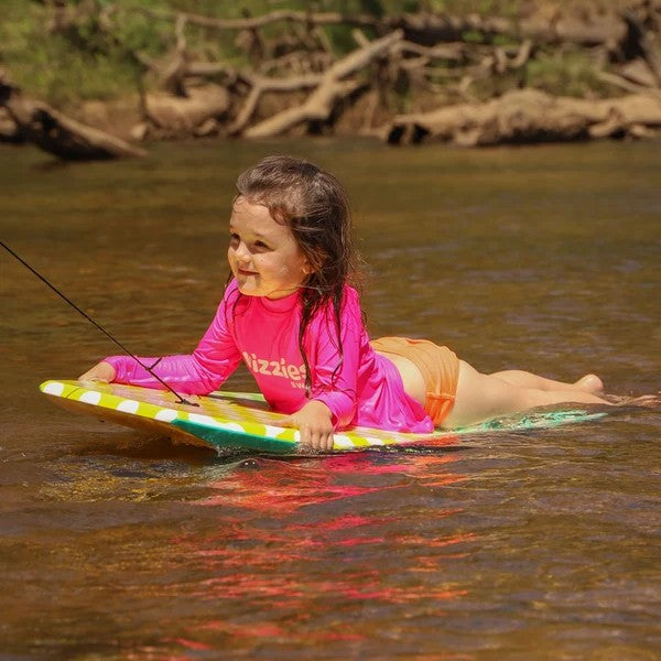 A young girl lies on her stomach on a colorful surfboard in a river, wearing a bright pink long-sleeve rash guard with 'Vizzies Swim' printed on the front and orange swim trunks.