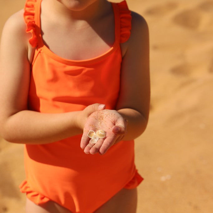 A child wearing a bright orange one-piece swimsuit from Vizzies Swim stands on a sandy beach, holding small seashells in their hand.