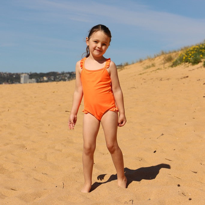 A young girl stands on a sandy beach wearing a bright orange one-piece swimsuit from Vizzies Swim with ruffled shoulder straps. The background features a clear blue sky, distant shoreline, and sand dunes.