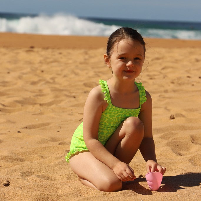 A young girl crouching on a sandy beach wearing a bright lime green one-piece swimsuit with a yellow pattern from Vizzies Swim. She is holding a small pink toy near the ocean waves in the background.