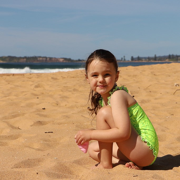 A young girl crouching on a sandy beach wearing a bright lime green one-piece swimsuit from Vizzies Swim, with the ocean and blue sky in the background.