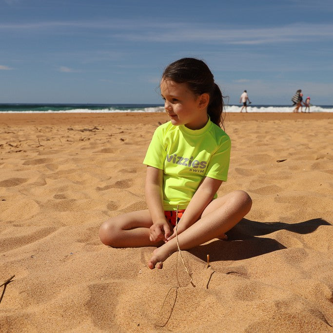 A young girl sits cross-legged on a sandy beach wearing a bright neon yellow rashie with 'Vizzies Swim' printed on the chest, with the ocean and blue sky in the background.
