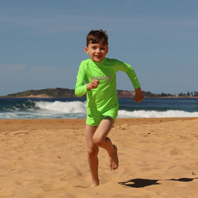 A young boy running on a sandy beach wearing a bright neon green long-sleeve swim shirt and matching swim trunks, with the ocean and blue sky in the background.