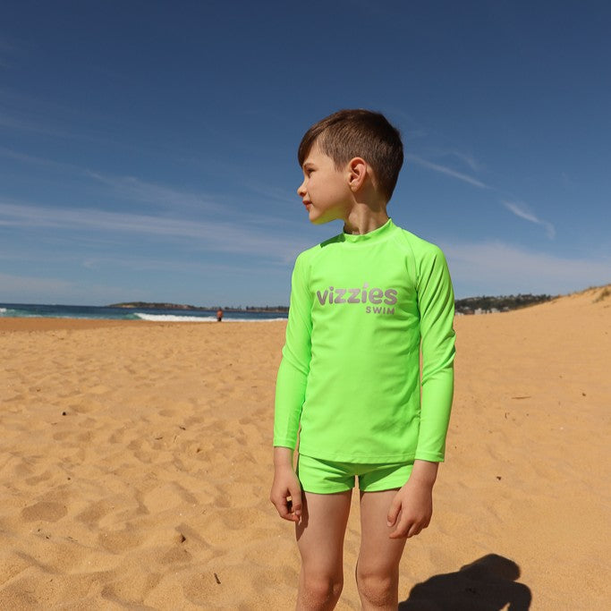 A young boy wearing a bright neon green long-sleeve rash guard and matching swim trunks stands on a sandy beach under a clear blue sky. The brand name 'vizzies swim' is printed on the chest of the rash guard.