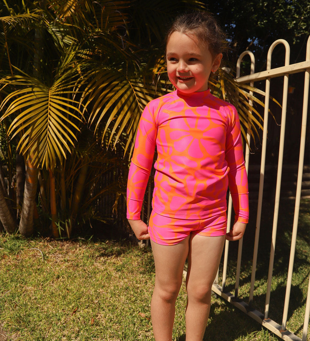 A young girl wearing a bright pink and orange patterned long-sleeve swim shirt and matching swim shorts, standing outdoors on grass in front of palm trees and a metal fence.