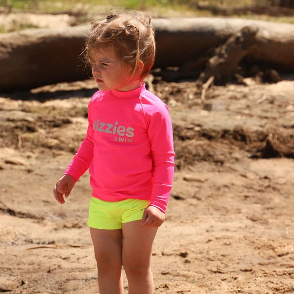 A young child standing outdoors on sandy ground wearing a bright pink long-sleeve rash guard with 'Vizzies Swim' printed on the chest and neon yellow shorts.
