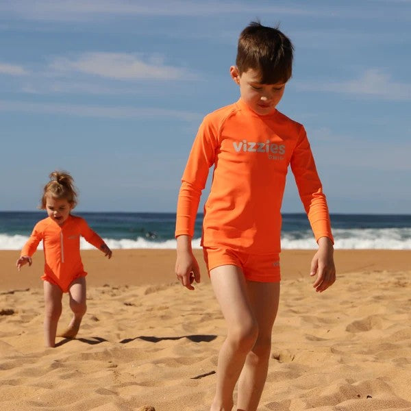 Two children wearing bright orange Vizzies swimwear outfits walking on a sandy beach with the ocean and blue sky in the background.