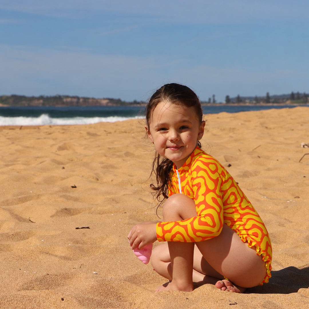 A young girl crouching on a sandy beach wearing a bright yellow and orange wavy long-sleeve swimsuit from Vizzies Swim, with the ocean and blue sky in the background.