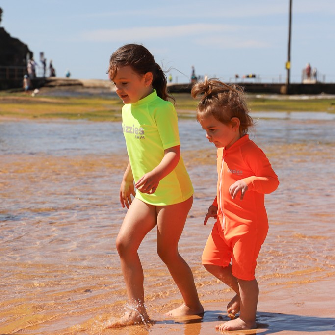 Two young children walking in shallow water at a beach. The older child wears a bright neon yellow short-sleeve rashie, and the younger child wears a bright orange swimsuit from Vizzies Swim.
