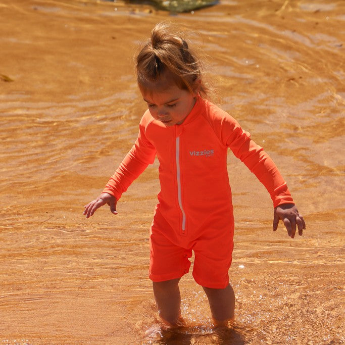 A toddler wearing a bright orange long-sleeve swim coverall with a front zipper and the brand name 'Vizzies Swim' printed on the chest, standing in shallow, sunlit water.