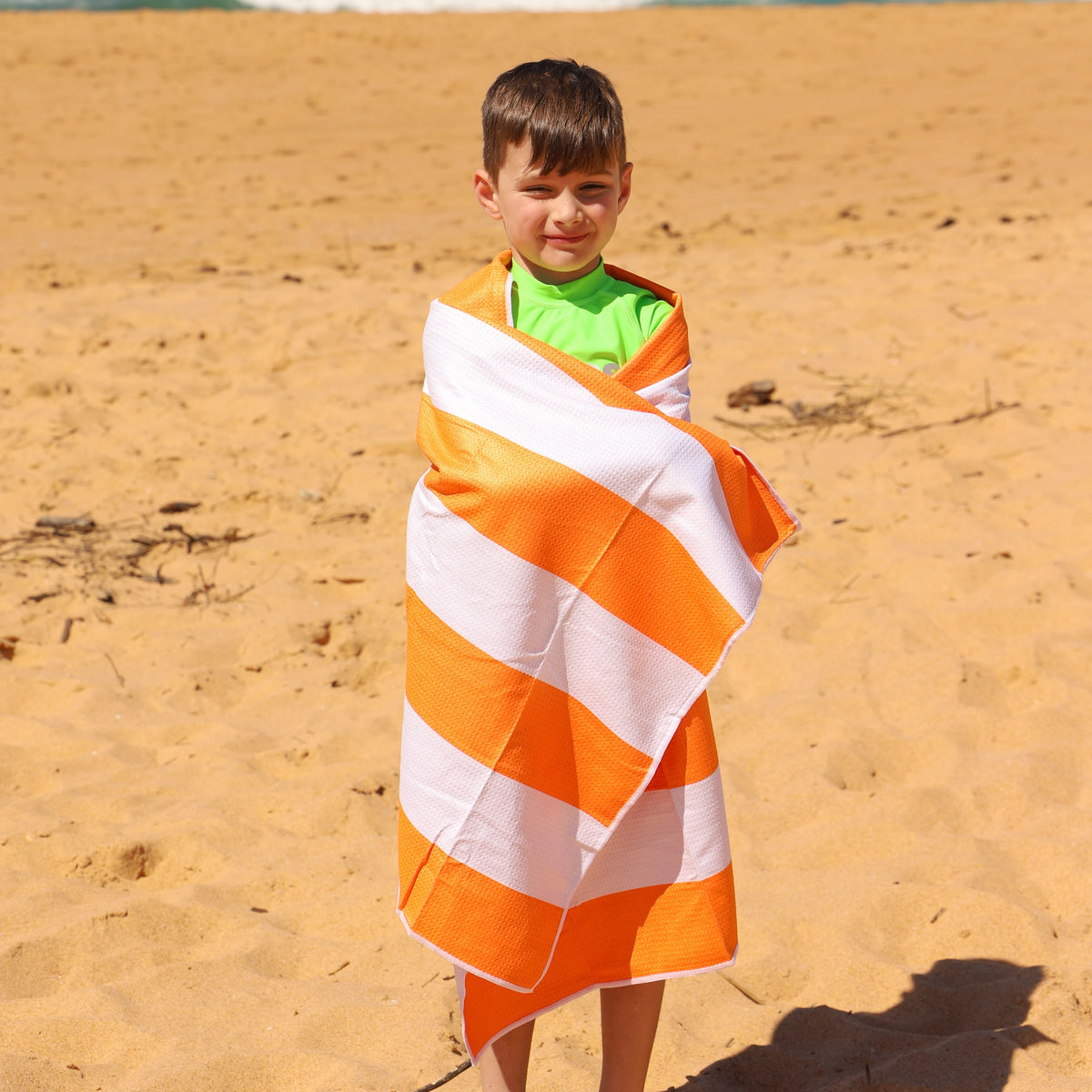 A young boy standing on a sandy beach wrapped in a Vizzies Swim striped orange and white beach towel, wearing a bright green Vizzies Swim rashie.