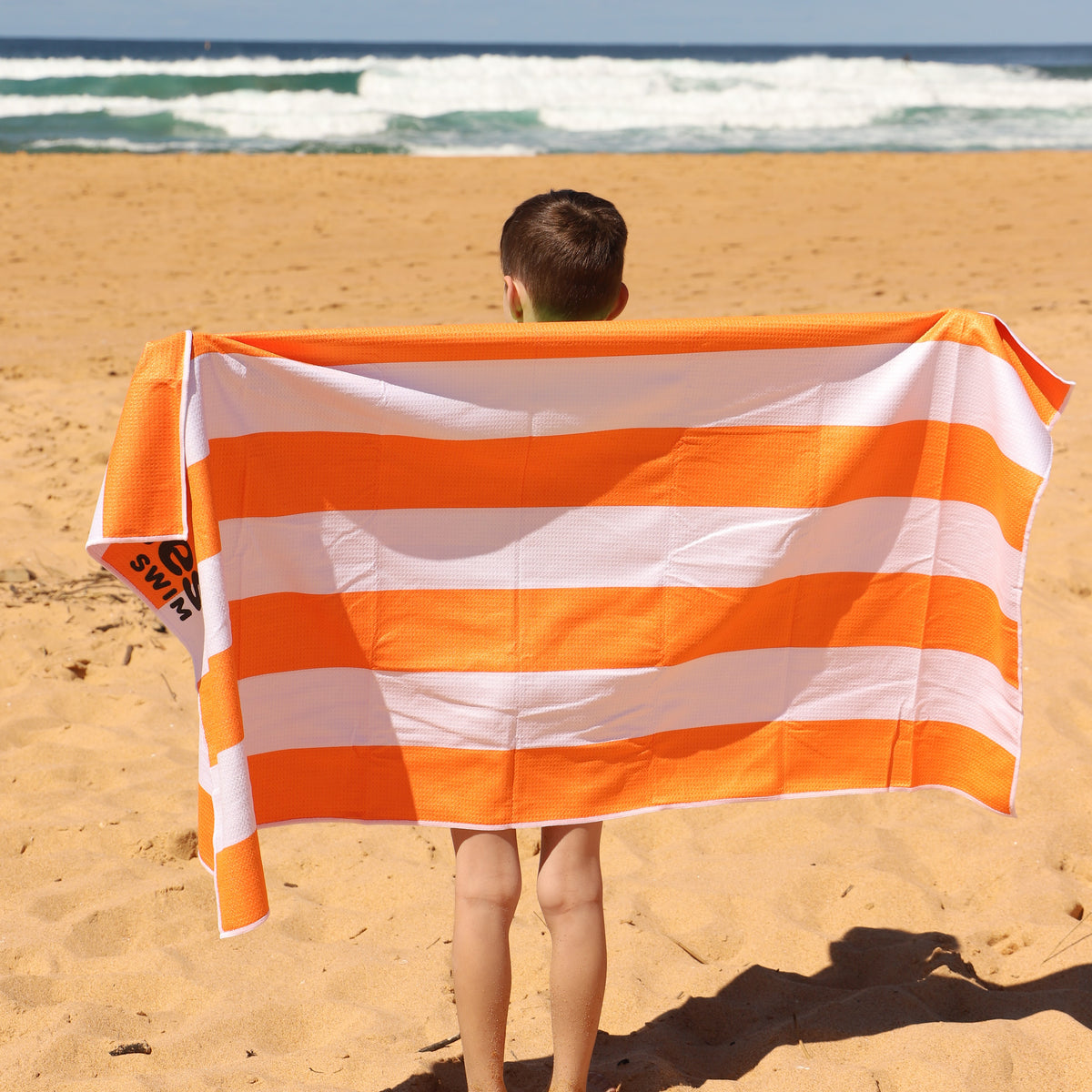 A child standing on a sandy beach with a Vizzies Swim orange and white striped beach towel spread behind them, with ocean waves in the background. 