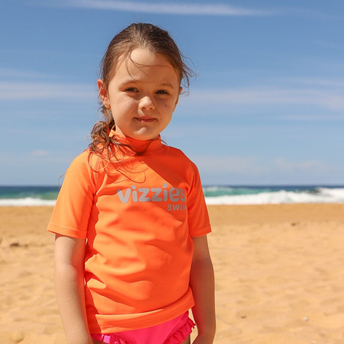 A young girl standing on a sandy beach wearing a bright orange short-sleeve rashie with 'Vizzies Swim' printed in light blue across the chest, with the ocean and blue sky in the background.