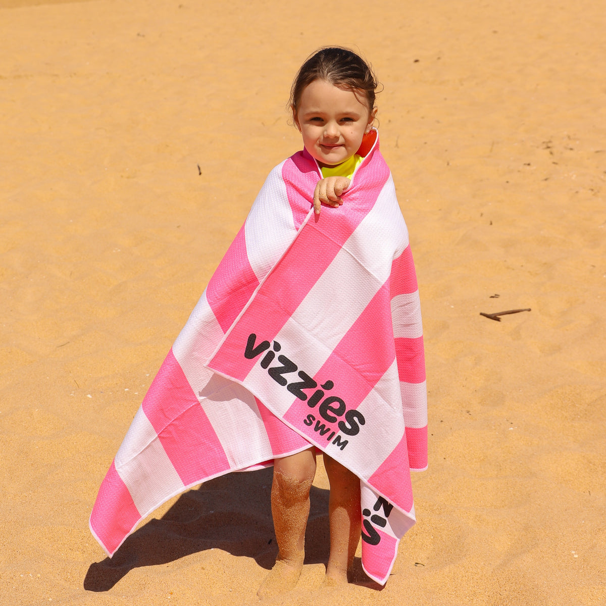 A young child standing on a sandy beach wrapped in a pink and white striped beach towel with the brand name 'Vizzies Swim' printed on it.