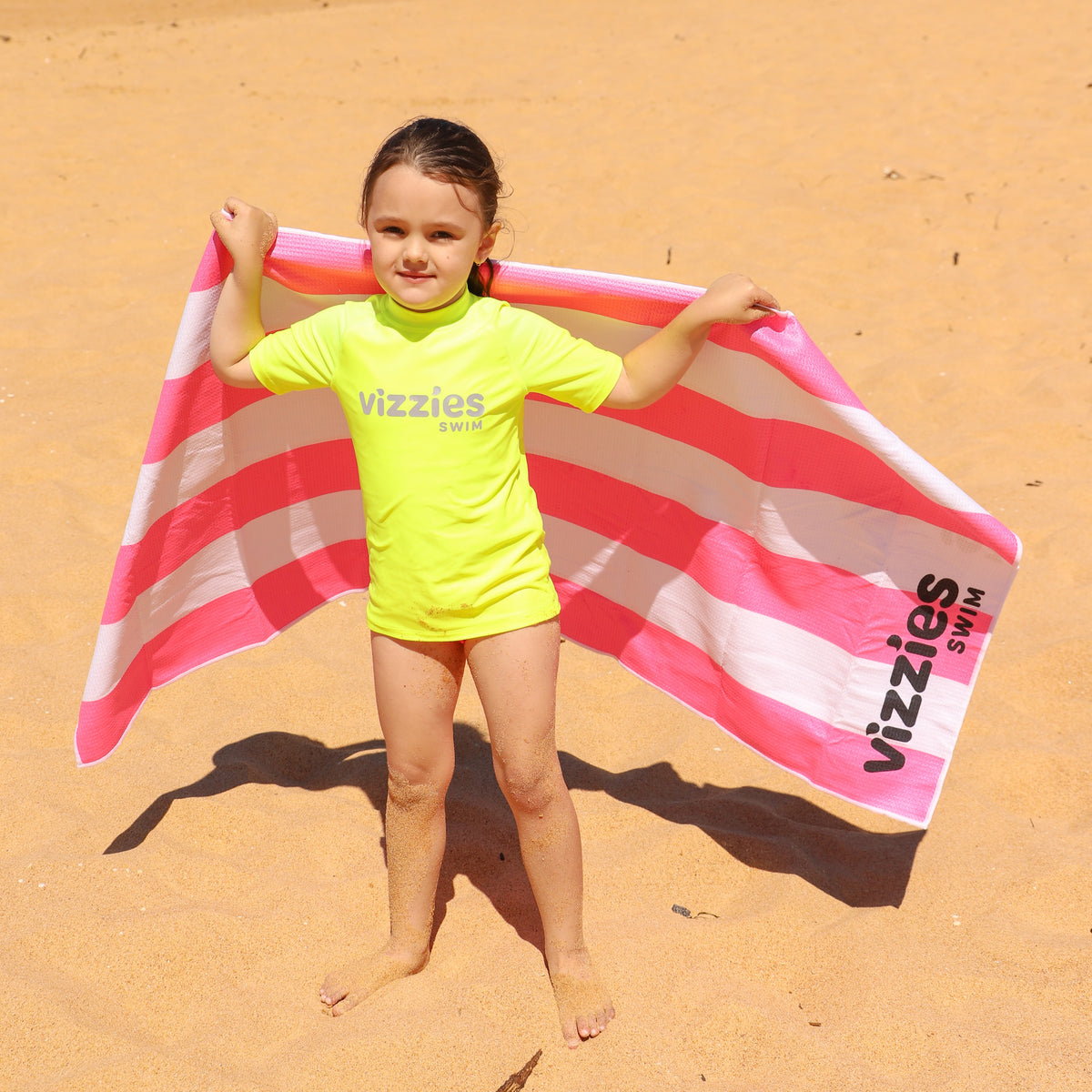 A young girl stands on a sandy beach holding up a large Vizzies Swim pink and white striped beach towel. She is wearing a neon yellow Vizzies swim short sleeve rashie.