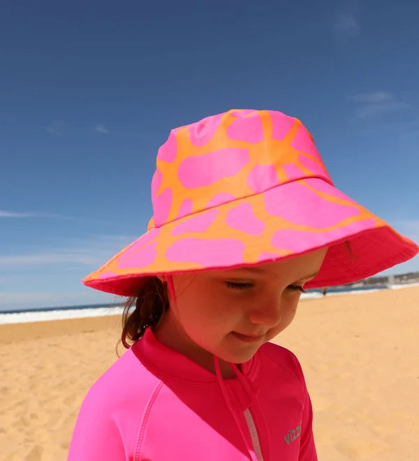 A young child wearing a bright pink wide-brim sun hat with an orange giraffe-like pattern, standing on a sandy beach under a clear blue sky.